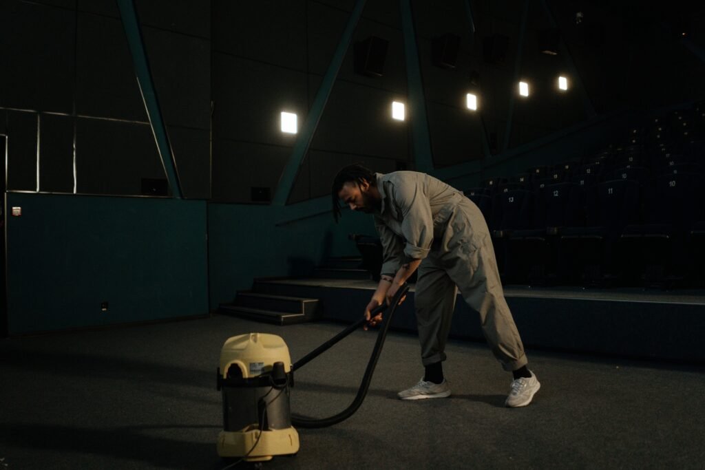 A cinema employee cleaning the theater floor with a vacuum cleaner under dim lights.